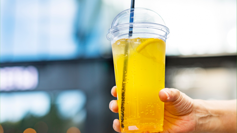Hand holds lemonade in a large plastic cup with a domed top and a black straw in it.