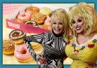 Dolly Parton and a Dolly Impersonator stand in front of a picture of her new Krispy Kreme doughnut line.